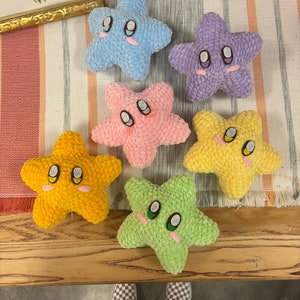May include: Six plush star-shaped toys in various colors: light blue, purple, pink, yellow, and green. Each star has embroidered eyes, rosy cheeks, and a smiling mouth. The toys are arranged on a striped fabric.