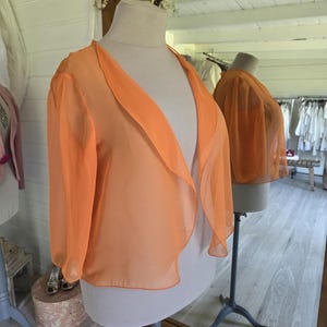 Peut inclure: Boléro orange vif en tissu transparent, présenté sur un mannequin. Ce vêtement à manches trois-quarts et devant ouvert est conçu pour être porté en superposition. La légèreté du tissu suggère une tenue estivale ou de soirée.