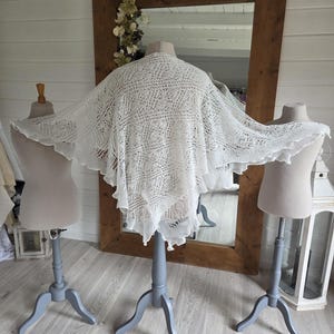 May include: A white, lacy shawl is draped over a mannequin. The shawl has a delicate, open weave design and a scalloped edge. The mannequin is on a gray stand with three legs. A large wooden framed mirror is in the background.