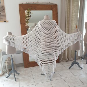 Puede incluir: Un chal blanco de crochet con un patrón triangular calado y flecos. El chal se exhibe sobre un maniquí, mostrando su diseño drapeado. El fondo incluye un espejo y otros elementos decorativos.