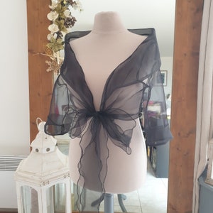 May include: A black sheer bolero-style wrap with three-quarter sleeves, displayed on a mannequin. The wrap is tied in a bow at the front, with flowing fabric cascading down. The background includes a mirror and decorative elements.