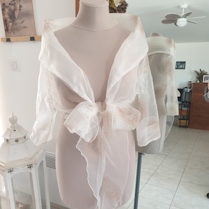 Può includere: Un scialle bianco in velo con un delicato motivo floreale dorato. Lo scialle è legato con un fiocco sul davanti e ha un design fluido ed elegante.