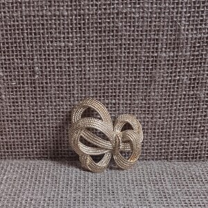 May include: A gold-toned brooch in a bow shape. The brooch is made of woven metal, with a textured appearance. The bow design features multiple loops and a central knot. The background is a textured, neutral-toned fabric.