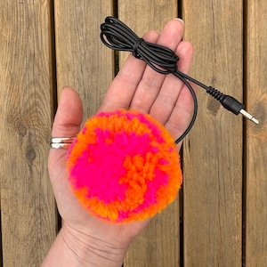 May include: A bright pink and orange pom-pom, attached to a black audio cable with a 3.5mm jack. The fluffy pom-pom is held in a hand, highlighting its vivid colours and handmade quality. A fun, decorative item.
