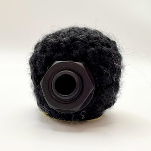 May include: A black, cylindrical component with a hexagonal base and a central aperture. It is encased in a dark, textured material, possibly yarn or fabric. The object rests on a light-coloured surface. It appears to be a part of a larger item or mechanism.