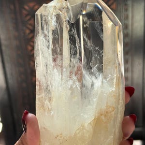 May include: A clear quartz crystal cluster with a natural, rough texture. The crystal has a pale yellow hue and is held in a hand. The crystal's facets reflect light, creating a sparkling effect. This is a natural gemstone.