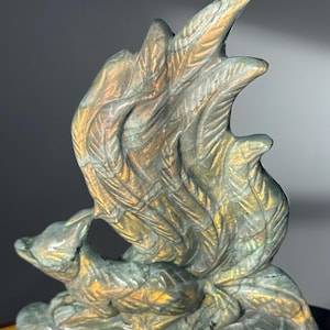 May include: A carved stone sculpture of a fox-like creature with a stylized wing or flame design. The sculpture has a metallic sheen with shades of gold, green, and gray. The fox is seated at the base of the sculpture.