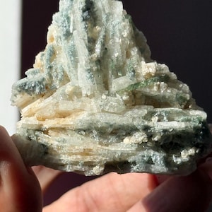 May include: A close-up of a mineral specimen featuring a cluster of light green, translucent crystals atop a layered, beige and green base. The crystals are vertically oriented, creating a textured, natural appearance.