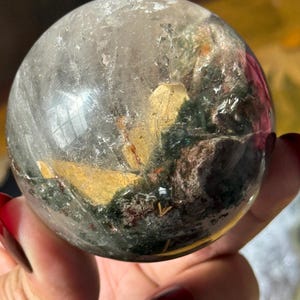 May include: A clear quartz crystal sphere with internal inclusions of green, gold, and brown minerals. The sphere is held in a hand with red nail polish. The crystal ball is a decorative item.
