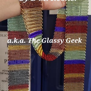 Op de afbeelding: Een lange sjaal gemaakt van in elkaar grijpende metalen ringen in een regenboogverloop van rood, oranje, geel, groen, blauw en violet. De sjaal bevat de tekst by D M. Rosner a.k.a. The Glassy Geek over het midden van de maliënkolderstof heen.