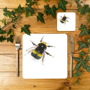 May include: A white ceramic coaster featuring a black and yellow bumblebee illustration. The coaster is placed on a wooden surface with green ivy.