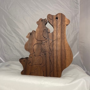 May include: A wooden puzzle featuring a bear family, with a large mother bear and several smaller bear cubs. The puzzle is made of dark brown wood, with visible wood grain. The bears are interlocked, creating a stacked design.