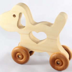 May include: A wooden toy dog with a heart-shaped cutout in its body. The dog is light-colored with brown wooden wheels. The toy is designed with a simple, clean aesthetic, suitable for young children. The dog is approximately 8 inches long.