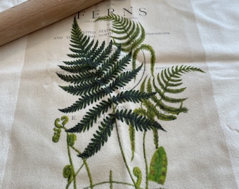 May include: A white tea towel with a green fern illustration and the word "FERNS" printed in black text. The tea towel is lying flat on a wooden surface with a wooden rolling pin on top of it.