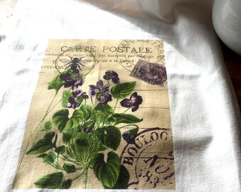 Op de afbeelding: Een witte keukendoek met een vintage Frans postkaartontwerp met paarse bloemen, groene bladeren en een bij. De postkaart heeft de tekst "Carte Postale" en een postzegel met de tekst "France".