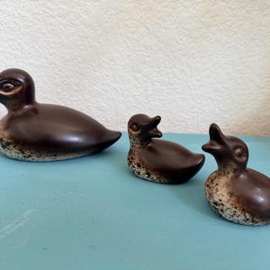 May include: A set of three ceramic duck figurines featuring a smooth dark brown finish on the upper bodies and a speckled cream and brown texture on the lower sections. The largest duck is positioned on the left, followed by two smaller ducklings.