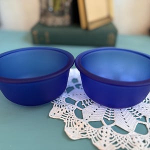 May include: Two cobalt blue glass bowls with a matte finish are displayed on a white lace doily. The bowls have a rounded shape with a dark blue rim. The background includes books and a light blue surface.