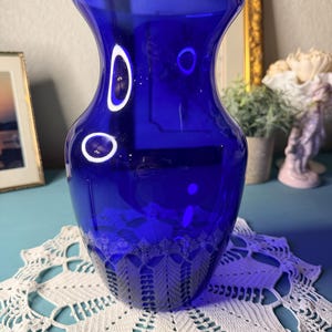 May include: A vibrant cobalt blue glass vase with a flared top and a decorative, etched pattern near the base. The vase sits on a white, crocheted doily. The background includes a framed picture and a gold-framed mirror.
