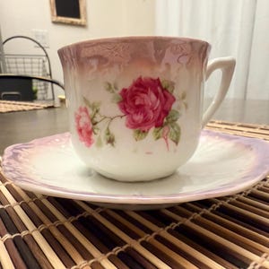 May include: A white teacup and saucer set with a pink rose floral design. The teacup has a handle and sits on a matching saucer. The set is placed on a bamboo mat.