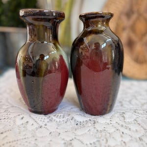 May include: Two small, glazed ceramic vases. The vases have a dark brown rim, transitioning to a deep red and green body. The vases are displayed on a white lace tablecloth.