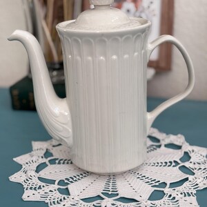 May include: A white ceramic teapot with a lid, a curved spout, and a handle. The teapot has vertical ribbed detailing and sits on a white lace doily. The background is a blurred image of a painting and some dried plants.