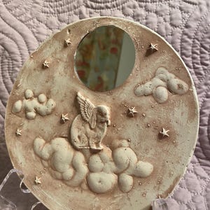 May include: A round, off-white decorative mirror with a sculpted design featuring a winged rabbit on clouds, surrounded by stars. The mirror has a small, circular reflective surface. The overall theme is whimsical and celestial.