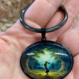 Puede incluir: Un llavero de metal negro con un colgante circular que presenta una escena de bosque con un conejo bajo la luna llena. El colgante tiene un acabado abovedado brillante. El llavero se sostiene en una mano.