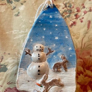May include: A hand-painted, winter-themed ornament. The blue background features white snowflakes. A snowman with a carrot nose and stick arms is in the center. Two brown rabbits are depicted, one holding a carrot. The ornament has a white string for hanging.