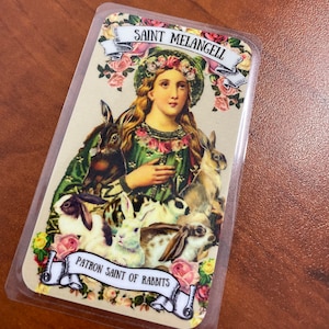 May include: A vintage-style religious card featuring a woman in a green dress and a crown of flowers. She is surrounded by rabbits and the text "Saint Melangell Patron Saint of Rabbits".