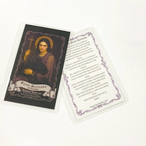May include: Two cards in clear plastic sleeves. One card features an illustration of Saint Gertrude of Nivelles holding a cross and a black cat. The other card has a prayer printed on it. The cards are on a white surface.