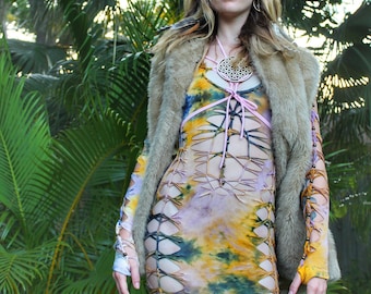 May include: A vibrant, tie-dye mini dress with cut-out detailing and long sleeves. The dress is worn with a faux fur vest and a decorative necklace. The background features lush green foliage and trees, creating a natural setting.