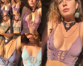 May include: A collection of five different coloured crochet tops with a criss-cross design at the front. The tops are all sleeveless and have a low-cut neckline. The model is wearing a variety of necklaces and earrings.