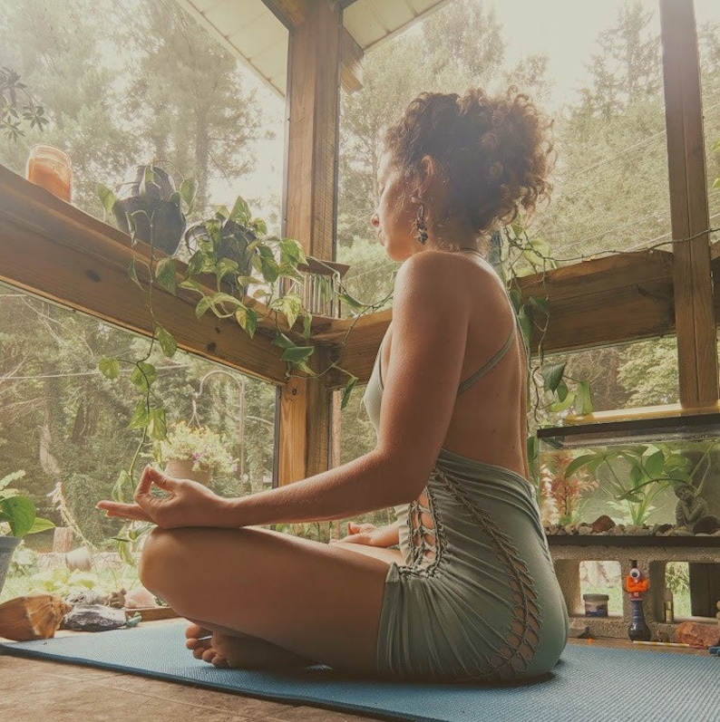May include: A woman sits in a yoga pose on a blue yoga mat in front of a window. She is wearing a light blue, sleeveless, lace-up dress. The window is open and there are plants and trees outside.