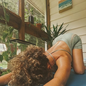 May include: A person with curly hair is performing a yoga pose on a blue mat. They are wearing a light blue tank top with a lace trim. The person is in a room with a window and plants.