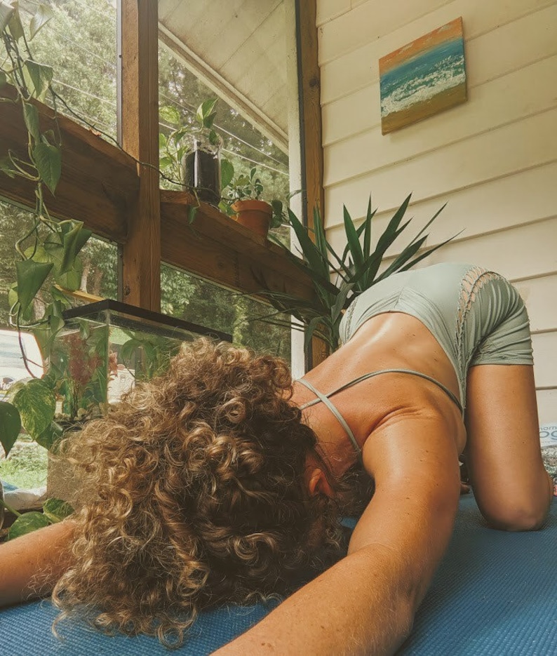 May include: A person with curly hair is performing a yoga pose on a blue mat. They are wearing a light blue tank top with a lace trim. The person is in a room with a window and plants.