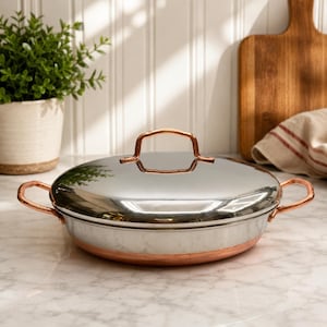 May include: A round, shallow cooking pan with a polished silver-tone lid and a copper-colored base. The pan features two side handles and a central lid handle, all finished in a matching copper tone, providing a dual-material aesthetic.