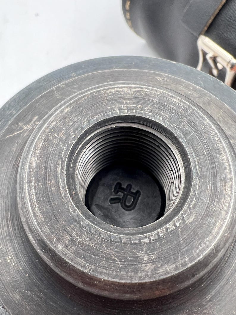 Pode incluir: Close-up de um componente met&aacute;lico cinza escuro com um orif&iacute;cio roscado central. O componente tem formato circular com cristas conc&ecirc;ntricas e uma marca&ccedil;&atilde;o "R" vis&iacute;vel dentro do orif&iacute;cio. O fundo inclui um vislumbre de um item de couro preto.