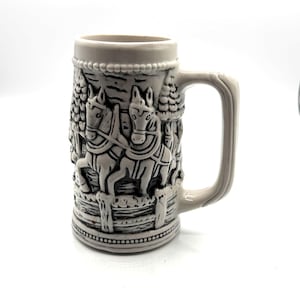 May include: A gray ceramic beer stein with a handle. The stein features a detailed relief of two horses pulling a carriage through a forest scene. The top and bottom rims are decorated with beaded designs.