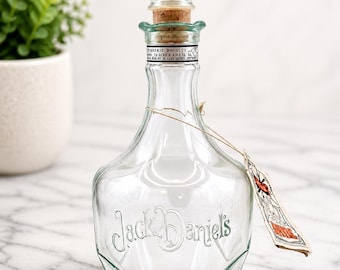 Puede incluir: Decantador de vidrio transparente con tapón decorativo y corcho. El decantador tiene el texto en relieve "Jack Daniel's" en la parte delantera. Una pequeña etiqueta está sujeta con una cuerda. El decantador está vacío y se encuentra sobre una superficie de mármol.