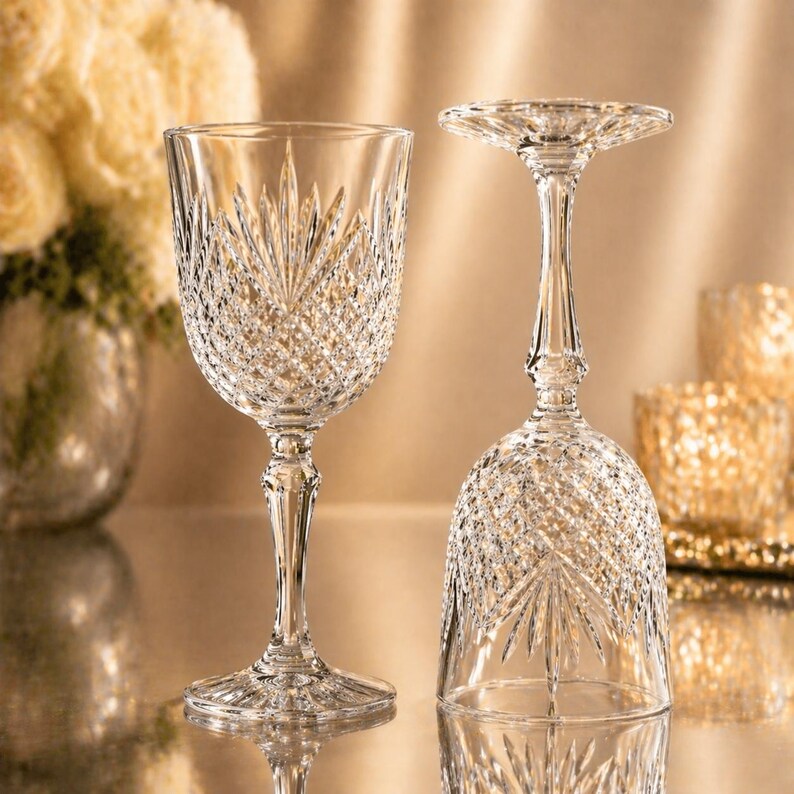 May include: Two clear crystal wine glasses with intricate cut glass patterns. One glass is upright, the other is inverted. The glasses have a detailed stem and a wide base. The background is a soft, golden hue.