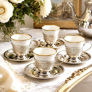 May include: A set of three white porcelain teacups with gold rims, each nestled in an ornate silver metal holder with a matching silver saucer. The intricate silver holders feature detailed embossed floral patterns and curved handles.