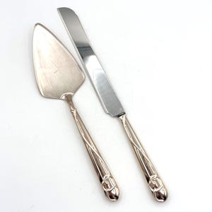 May include: Silver cake server and knife set. The server has a triangular blade, and the knife has a serrated edge. Both have decorative handles with a floral design. The set is made of a shiny, reflective metal.