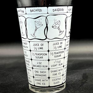 May include: A clear cocktail mixing glass with a black rim and a white and light blue design. The glass features cocktail names and ingredient lists, including "Bacardi" and "Daiquiri". The design includes illustrations of cocktail glasses.