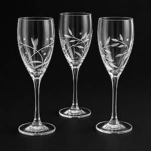 May include: Three clear glass wine glasses with slender stems and circular bases. Each glass features a delicate, etched pattern of thin, winding vines with small leaves and scattered dots encircling the upper portion of the bowl.