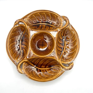 May include: A brown ceramic leaf-shaped serving dish with a central circular well. The dish has four large, detailed leaf designs around the perimeter, with a white accent in the crevices. The dish is on a white background.