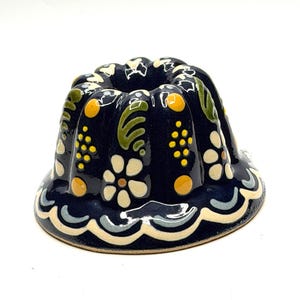 May include: A dark blue ceramic bundt cake mold featuring a hand-painted floral pattern with white daisies, green leaves, and yellow dots. The fluted sides have a scalloped base edge with light blue and cream accents, creating a decorative finish.