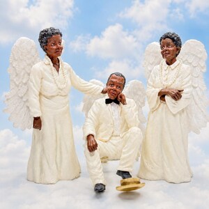 May include: Three angel figurines with white wings and cream-colored robes stand against a blue sky background. One figure is seated, wearing a suit and bow tie, with a hat on the ground. The other two figures are standing.