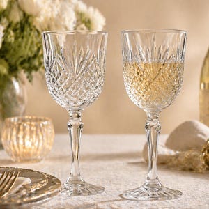 May include: Two crystal wine glasses with intricate diamond-cut patterns, one filled with a golden liquid. The glasses sit on a table with a floral arrangement and a lit candle in the background. A bottle of wine is also visible.