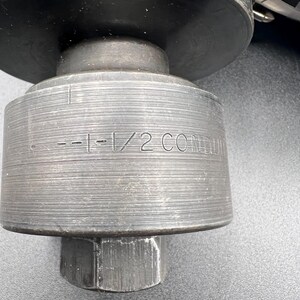 Pode incluir: Close-up de uma ferramenta de metal cinza escuro e resistente. A ferramenta tem um corpo cil&iacute;ndrico com o texto "--1-1/2 CONDUIT" gravado na lateral. Uma porca sextavada &eacute; vis&iacute;vel na base. A ferramenta parece ser para uso industrial.