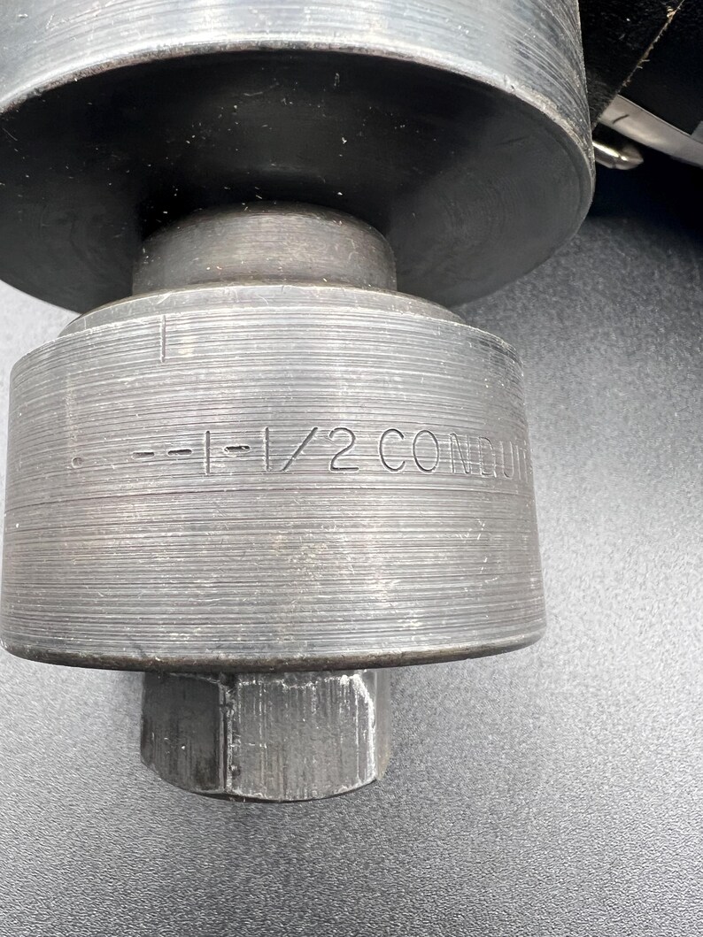 Pode incluir: Close-up de uma ferramenta de metal cinza escuro e resistente. A ferramenta tem um corpo cil&iacute;ndrico com o texto "--1-1/2 CONDUIT" gravado na lateral. Uma porca sextavada &eacute; vis&iacute;vel na base. A ferramenta parece ser para uso industrial.